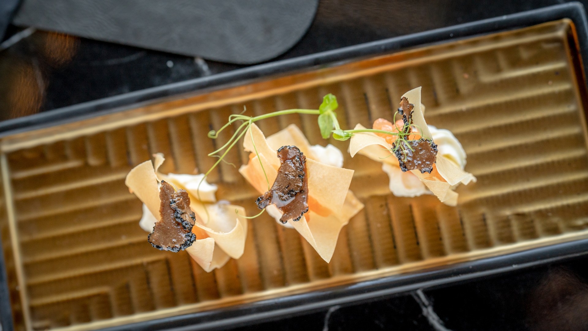 Tre fagottini di sfoglia con tartufo nero a lamelle, uova di salmone e germogli su vassoio dorato rigato di Inari Sushi