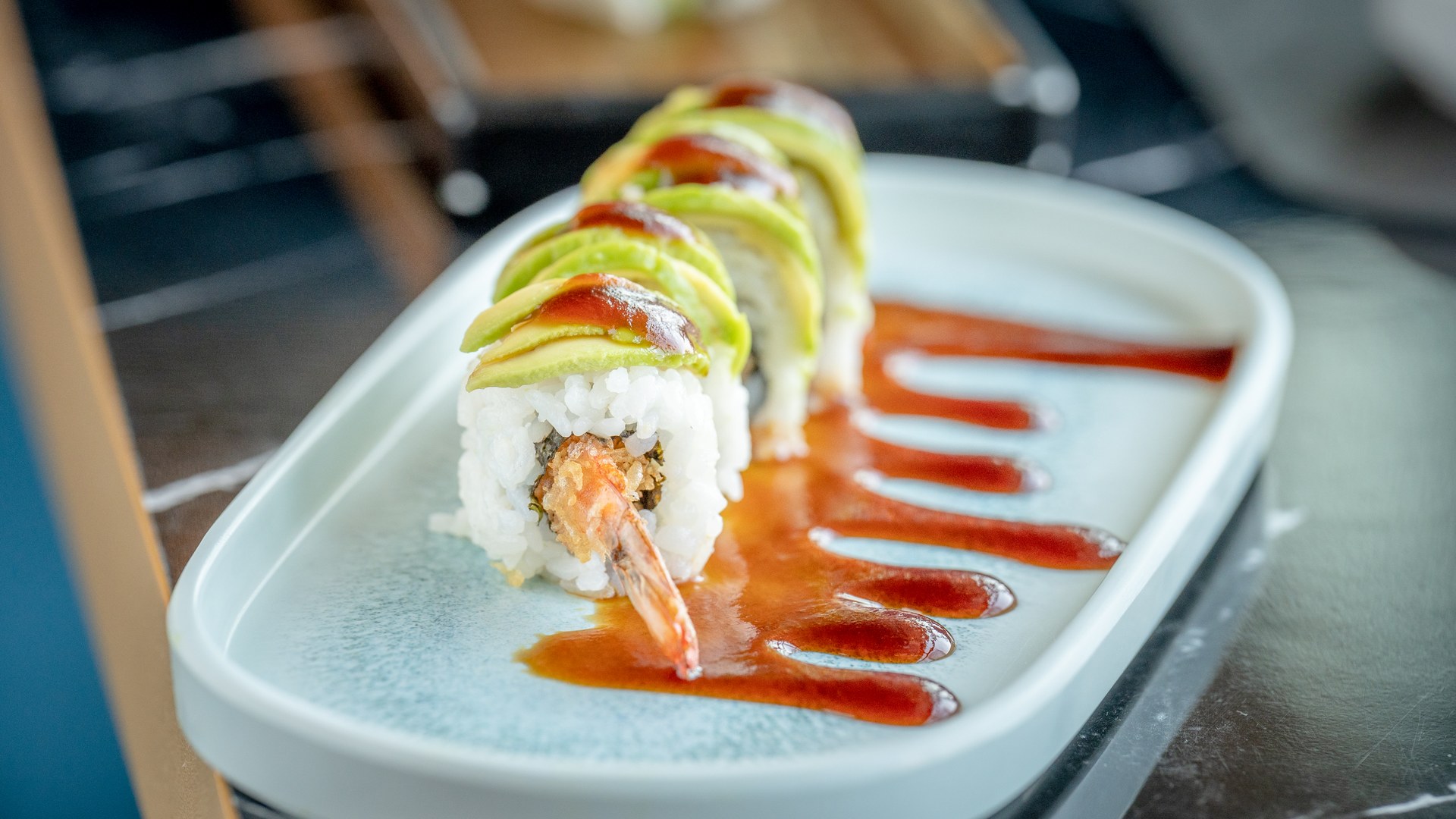 Uramaki con gambero fritto e fettine di avocado, salsa ambrata su piatto ovale celeste di Inari Sushi